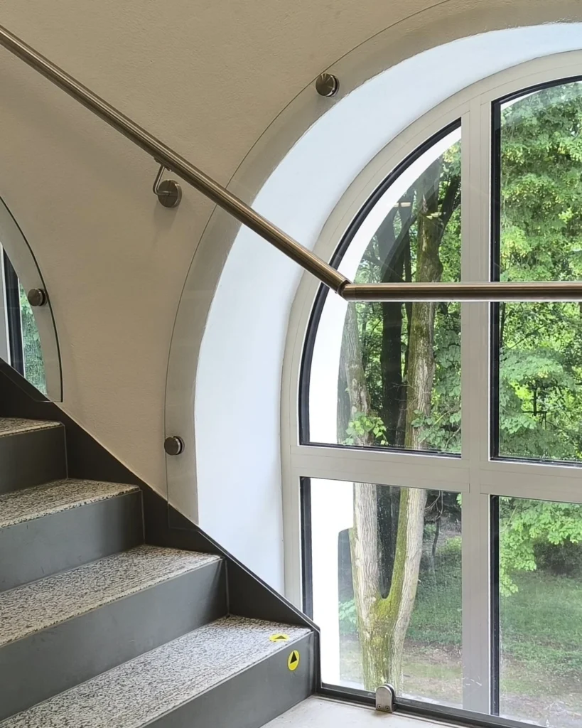 Detail uchytenia skleneného zábradlia na schodisku pomocou nerezových terčov priamo do steny okolo oblúkového výrezu okna.